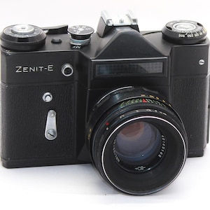 May include: A black Zenit-E 35mm film camera featuring a textured body, a prominent lens with visible aperture and focus rings, a top-mounted shutter speed dial, a film advance lever, and a hot shoe mount for external flash attachments.