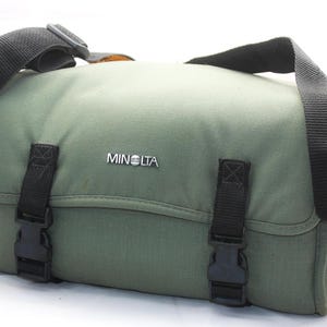 May include: An olive green camera bag featuring a black adjustable shoulder strap and two black plastic buckle closures on the front flap. The word MINOLTA is printed in white letters on a small black rectangular badge centered on the front flap.