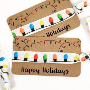 May include: Two white elastic hair ties with a Christmas light string design. The hair ties are wrapped around brown cards with the text "Happy Holidays".