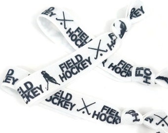 Könnte beinhalten: Drei weiße Gummibänder für Haare mit dem Text "FIELD HOCKEY" und einer Stickfigur, die Feldhockey spielt, in Schwarz gedruckt.