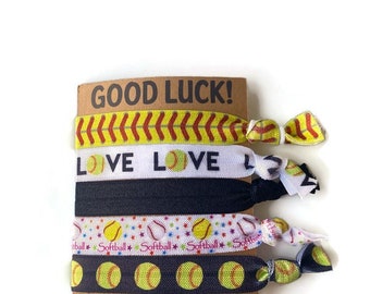 May include: Set of five softball-themed hair ties. The hair ties feature different designs, including a softball with red stitching, a softball with stars, and a softball with the words "Love Love". The hair ties are packaged on a brown card with the words "Good Luck!" printed on it.