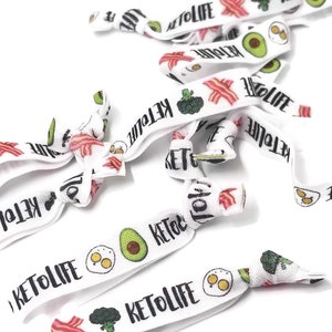 May include: White elastic hair ties with a keto-themed design. The hair ties feature images of avocado, bacon, eggs, and broccoli. The text "Keto Life" is printed on the hair ties.