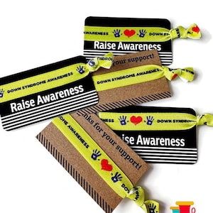 Puede incluir: Cuatro pulseras amarillas y verdes con el texto "Down Syndrome Awareness" y un corazón rojo con dos manos azules. Las pulseras están en tarjetas negras y marrones con el texto "Raise Awareness" y "¡Gracias por tu apoyo!"