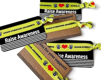 May include: Four yellow and green wristbands with the text "Down Syndrome Awareness" and a red heart with two blue hands. The wristbands are on black and brown cards with the text "Raise Awareness" and "Thanks for your support!"