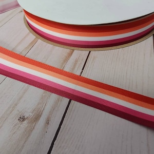 May include: A close-up of a lesbian pride ribbon with red, orange, yellow, white, pink, and dark pink stripes. The ribbon is made of satin and is lying on a wooden surface.