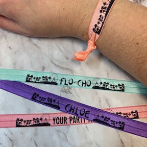 May include: Three colorful elastic bracelets with a Ferris wheel and palm tree design. The bracelets are labeled "Flo-Cho", "Chloe", and "Your Party".