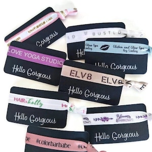 May include: A collection of colourful hair ties with various designs and text on black cards. The cards have the text "Hello Gorgeous" printed on them. The hair ties include designs such as "Love Yoga Studio", "Glow Spa", "Glisten and Glow Spa", "Hair Holly", "#colorbarbabe", and "Blooms & Arrows".