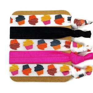 May include: Set of five colorful hair ties with a cupcake pattern. The hair ties are on a brown card. Two of the hair ties are black, one is pink, and two are white with a cupcake pattern.
