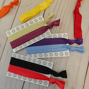 May include: Assortment of colorful elastic hair ties in various colors, including red, orange, yellow, gold, purple, blue, and black. Each tie is knotted at the ends. The ties are displayed on white cards with volleyball graphics.