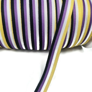 Pode incluir: Um rolo de fita listrada de roxo, branco, preto e amarelo. A fita tem aproximadamente 1 polegada de largura.