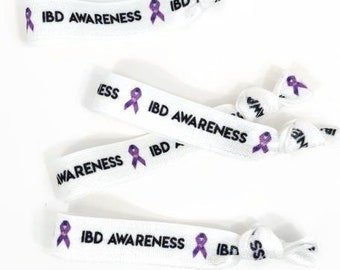 Op de afbeelding: Vijf witte elastische polsbandjes met een paars lint en de tekst "IBD AWARENESS" erop gedrukt.