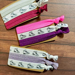 Op de afbeelding: Een verzameling elastische haarbanden in verschillende kleuren, waaronder roze, paars en bordeauxrood. Elke band heeft een herhaald patroon van schaatsen en is bevestigd aan een kleine kartonnen achterkant. De uiteinden van de banden zijn geknoopt.