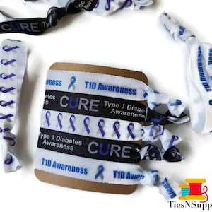 May include: A set of six white elastic hair ties with blue and black ribbon accents. The ribbon features the text "T1D Awareness", "Type 1 Diabetes Awareness", and "CURE".