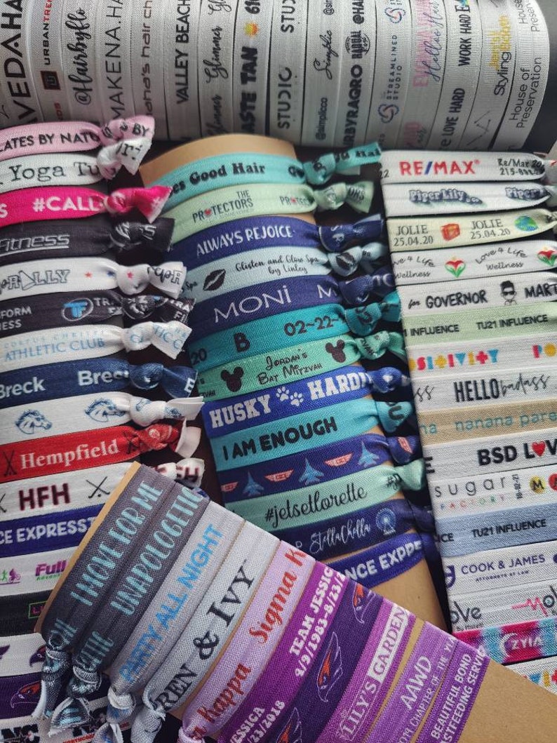 May include: A close-up of a variety of colourful hair ties with different designs and text. The hair ties are made of elastic material and have a variety of different colours, including blue, purple, pink, green, and white. The text on the hair ties includes phrases such as "I am enough", "Always Rejoice", "Husky", "Team Jessica", "Lily's Garden", and "RE/MAX".