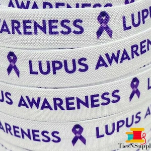 Könnte beinhalten: Weißes Band mit dem Text "Lupus Awareness" in lila gedruckt. Das Band ist mit einem lilafarbenen Sensibilisierungsband verziert.