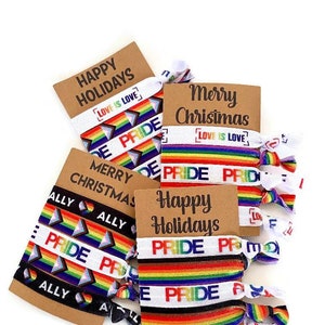 May include: A set of colorful rainbow pride hair ties with the words "Happy Holidays", "Merry Christmas", "Love is Love", and "Ally" printed on them. The hair ties are packaged on brown cards.