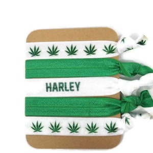 Könnte beinhalten: Drei weiße Gummibänder für Haare mit grünen Marihuana-Blatt-Designs. Das mittlere Gummiband hat den Namen "HARLEY" in Grün gedruckt.