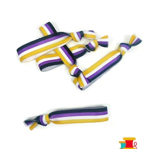 Pode incluir: Um conjunto de quatro elásticos de cabelo listrados em roxo, branco, amarelo e preto. Os elásticos de cabelo são feitos de material elástico e estão amarrados em um nó.
