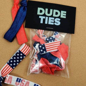 May include: A clear plastic bag filled with red, white, and blue elastic hair ties with a black card that says "DUDE TIES" on it. The hair ties have an American flag design.