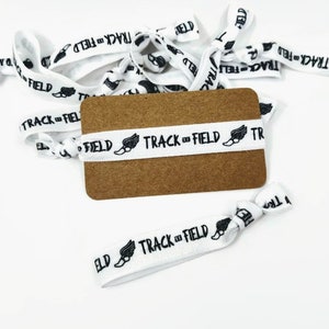 May include: White elastic hair ties with the text "TRACK & FIELD" and a running shoe icon printed on them. The hair ties are bundled together on a brown card.