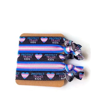 May include: A set of four black elastic hair ties with a white, light blue, and pink striped pattern and a transgender pride heart with the text "Protect Trans Kids" repeated on each tie.