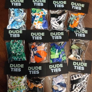 May include: Twelve small clear plastic bags with black labels that say "DUDE TIES" and contain colorful hair ties with various patterns. The patterns include: green shamrocks, American flags, orange and blue stripes, green and blue tie-dye, black and white skulls, avocado, and more.