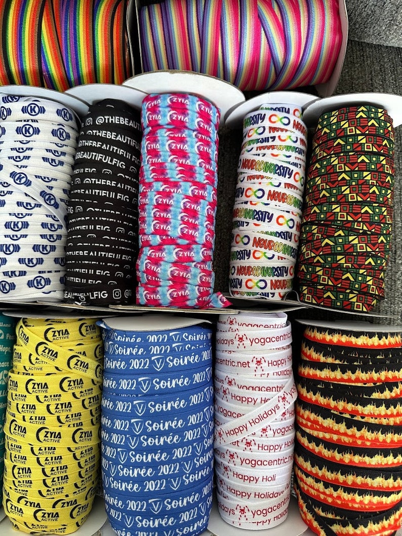 Peut inclure: Une collection d'&eacute;lastiques &agrave; cheveux color&eacute;s avec diff&eacute;rents motifs et textes. Les motifs comprennent des rayures arc-en-ciel, un motif tie-dye, un motif noir et blanc avec le texte "Neurodiversity", un motif noir et vert avec des formes g&eacute;om&eacute;triques, un motif jaune et noir avec le texte "Ziya Active", un motif bleu et blanc avec le texte "Soir&eacute;e 2022", un motif blanc et rouge avec le texte "Happy Holidays", et un motif noir et orange avec des flammes.