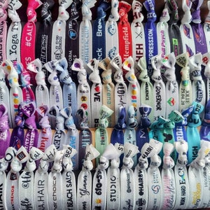 May include: A collection of colourful hair ties with various text and designs. The hair ties are made of elastic material and have a variety of colours, including white, blue, pink, purple, and green. The text on the hair ties includes names, phrases, and logos.