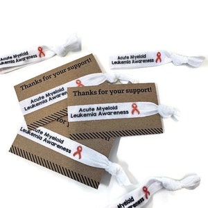May include: White elastic hair ties with the text "Acute Myeloid Leukemia Awareness" and an orange ribbon printed on them. The hair ties are displayed on brown cards with the text "Thanks for your support!" printed on them.