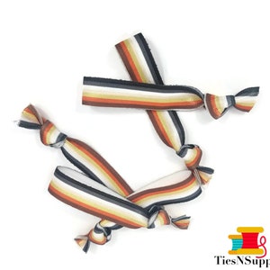 Pode incluir: Três elásticos de cabelo listrados em marrom, preto, branco e dourado. Os elásticos de cabelo estão amarrados em nós.