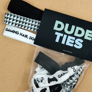 May include: A package of black and white hair ties with the text "DUDE TIES" on the front. The hair ties have different designs, including a geometric pattern, a video game controller, and the text "DON'T CARE".