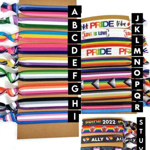 May include: Assortment of colorful woven bracelets with various pride-themed designs, including rainbow stripes, the words "Pride Tribe", "Love is Love", and "Pride 2022", and the phrase "Protect Trans Kids".