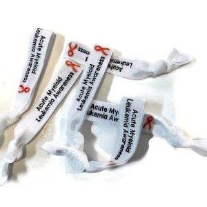 Peut inclure: Des élastiques à cheveux blancs avec le texte "Acute Myeloid Leukemia Awareness" et un ruban orange imprimé dessus.