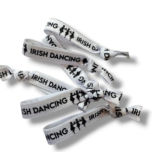 Op de afbeelding: Witte elastische haarbandjes met zwarte tekst "Irish Dancing" en een silhouet van dansers.