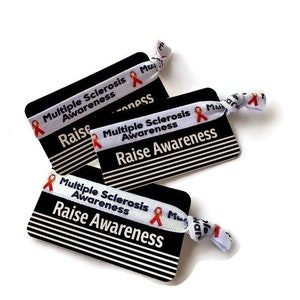 May include: Three white elastic wristbands with the text "Multiple Sclerosis Awareness" and a red ribbon printed on them. The wristbands are tied together and are on top of three black and white cards with the same text and red ribbon printed on them. The cards have the text "Raise Awareness" printed on them.