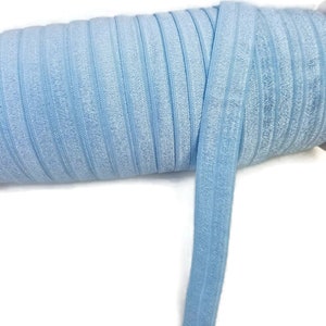 May include: A roll of light blue elastic, also known as fold over elastic or FOE, is shown on a white background. The elastic is folded and rolled up, with a small portion of the elastic unrolled to show the texture.