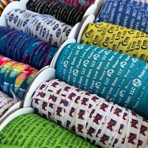 Peut inclure: Gros plan sur des bobines de ruban color&eacute; avec diff&eacute;rents motifs et textes. Le texte sur les rubans comprend "Peace Love Hope, LLC", "Spin Hair Don't Care" et "Zia Active".