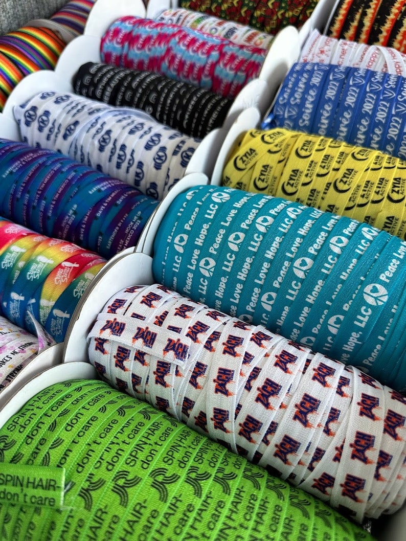 Peut inclure: Gros plan sur des bobines de ruban color&eacute; avec diff&eacute;rents motifs et textes. Le texte sur les rubans comprend "Peace Love Hope, LLC", "Spin Hair Don't Care" et "Zia Active".
