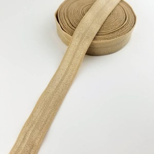 May include: A roll of beige elastic trim with a satin finish. The elastic is rolled up and a length of the trim is extending out.