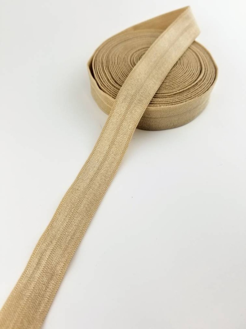 May include: A roll of beige elastic trim with a satin finish. The elastic is rolled up and a length of the trim is extending out.