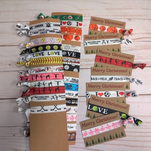 May include: A set of colorful hair ties with various sports and holiday themes. The hair ties are arranged on a brown card with the text "Merry Christmas!" on each card. The hair ties feature designs such as basketballs, baseballs, soccer balls, golf balls, cheerleaders, gymnasts, and track and field.