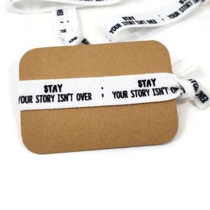 Op de afbeelding: Witte elastische haarbandjes met de tekst "Stay Your Story Isn't Over" in zwart gedrukt. De haarbandjes worden getoond op een bruine kaart.
