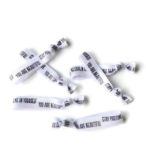 May include: White elastic hair ties with black text. The text on the hair ties reads: "You are beautiful", "Stay Positive", "Believe in yourself", "You are beautiful", "Stay Positive", and "You are beautiful".