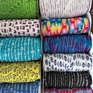Peut inclure: Une collection d'&eacute;lastiques &agrave; cheveux color&eacute;s avec diff&eacute;rents motifs et textes. Les &eacute;lastiques sont dispos&eacute;s en rang&eacute;es sur une surface blanche. Certains des motifs incluent un motif arc-en-ciel, un signe de paix et le texte "Soir&eacute;e 2022".