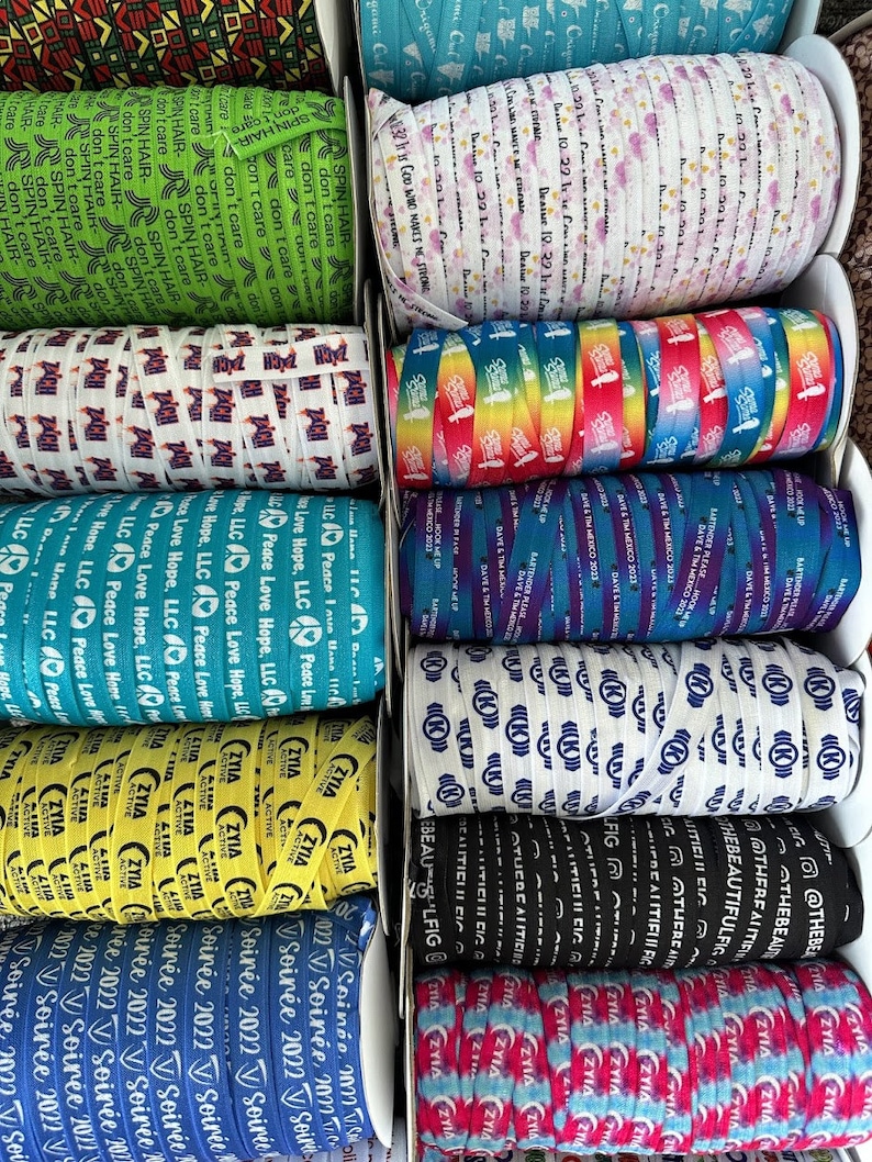 Peut inclure: Une collection d'&eacute;lastiques &agrave; cheveux color&eacute;s avec diff&eacute;rents motifs et textes. Les &eacute;lastiques sont dispos&eacute;s en rang&eacute;es sur une surface blanche. Certains des motifs incluent un motif arc-en-ciel, un signe de paix et le texte "Soir&eacute;e 2022".