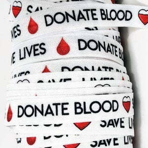 Puede incluir: Un rollo de cinta elástica blanca con el texto "Donar sangre Salva vidas" impreso en negro. La cinta está decorada con corazones rojos e iconos de gotas de sangre.