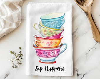 May include: White tea towel featuring a stack of five colorful teacups with floral designs. The teacups are in shades of blue, yellow, pink, and orange. The text "Sip Happens" is printed in black script at the bottom of the towel.
