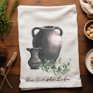 May include: White kitchen towel with a watercolor illustration of two gray vases and green foliage. The text "The Simple Life" is printed in black script at the bottom. The towel is displayed on a wooden surface.