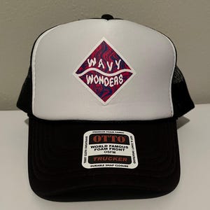 Puede incluir: Una gorra de camionero negra y blanca con un gráfico en forma de diamante con patrones ondulados rojos y azules y el texto Wavy Wonders. El panel frontal es de espuma blanca, mientras que la visera y la malla trasera son negras. Etiqueta Otto.