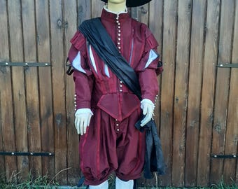 May include: A maroon and white patterned doublet with a black sash, worn with maroon breeches and white boots. The outfit is topped with a black feathered hat.